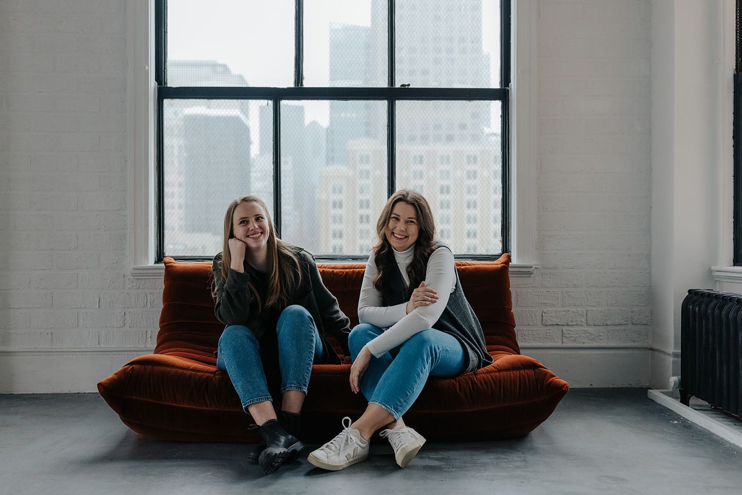 Two friends relax on an orange couch in a minimalist urban loft space.