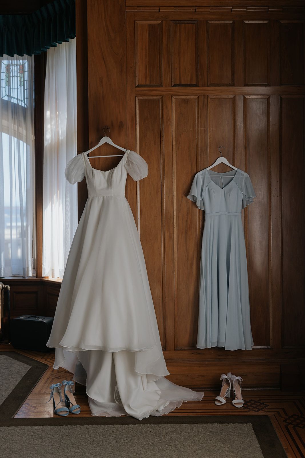 Wedding attire displayed on hangers against wooden paneled walls including a white gown and blue bridesmaid dress.