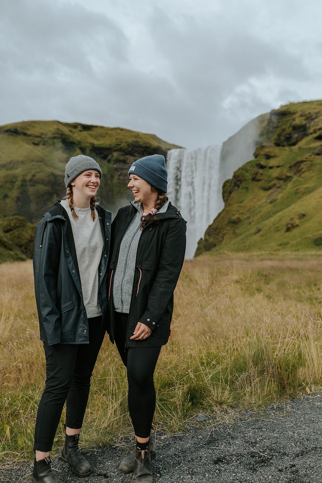 Friends sharing laughter in front of a waterfall on a cloudy day in Iceland wearing matching winter gear.
