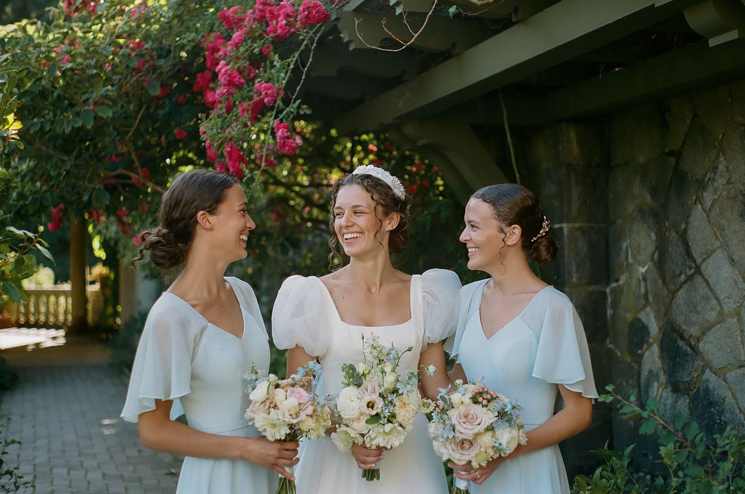 Three women in white dresses share joyful laughter while holding bouquets.