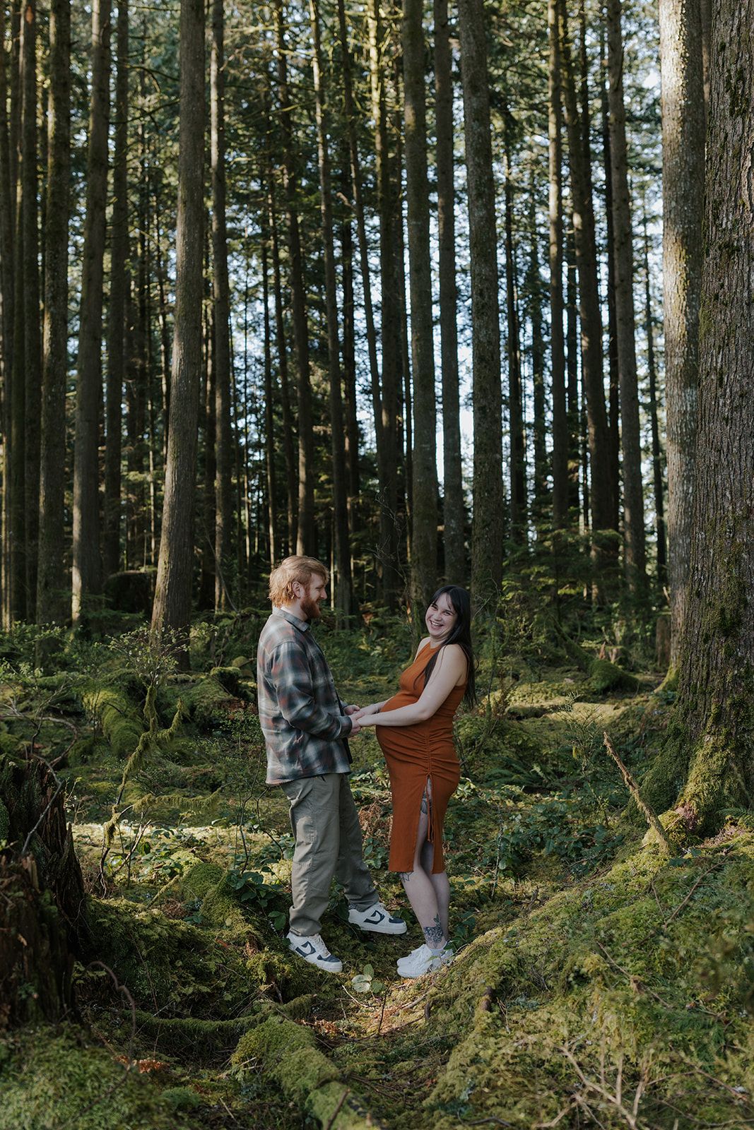 Couple standing together on mossy ground in a dense evergreen forest.