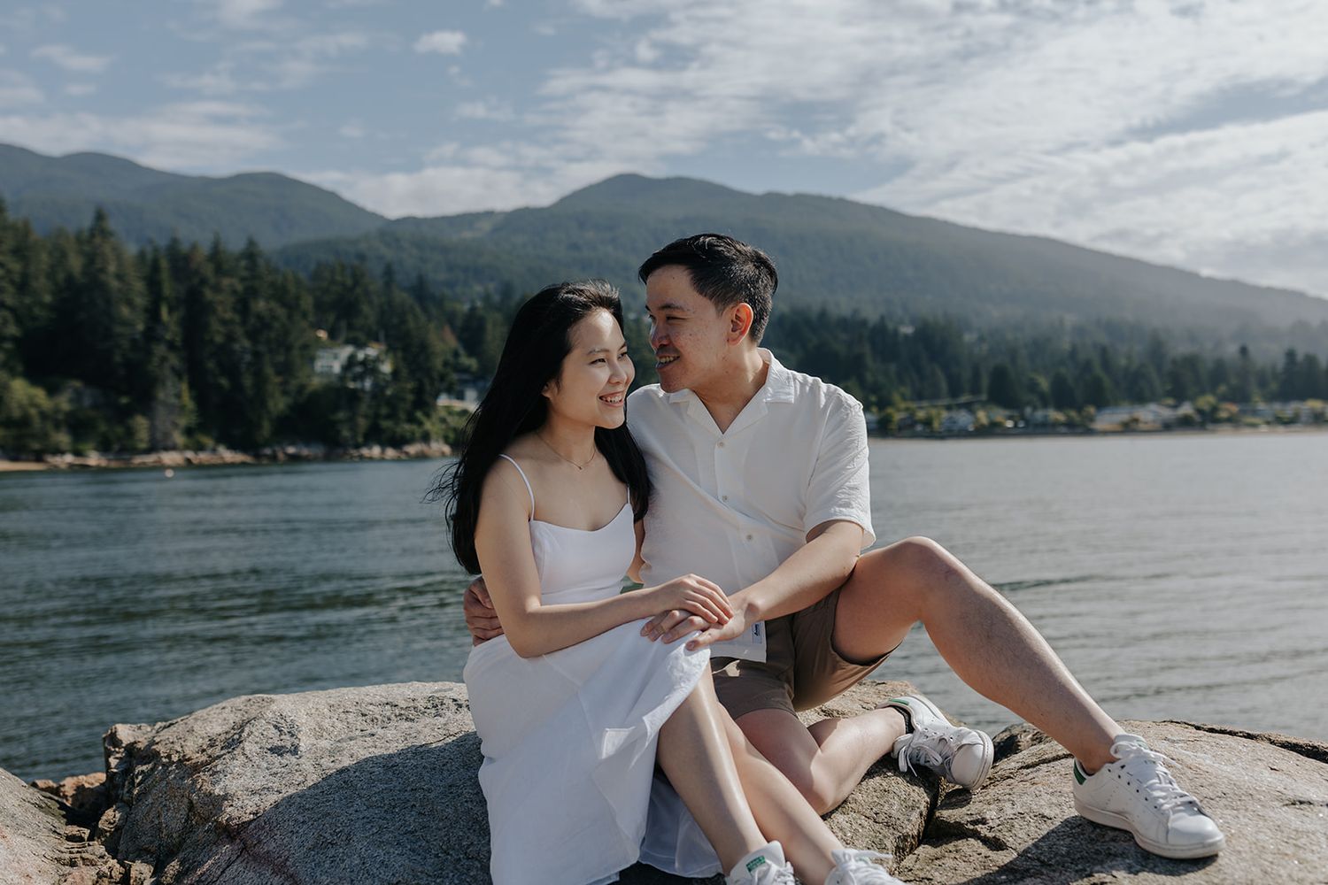A couple sits together on a beach with mountains and water in the background.