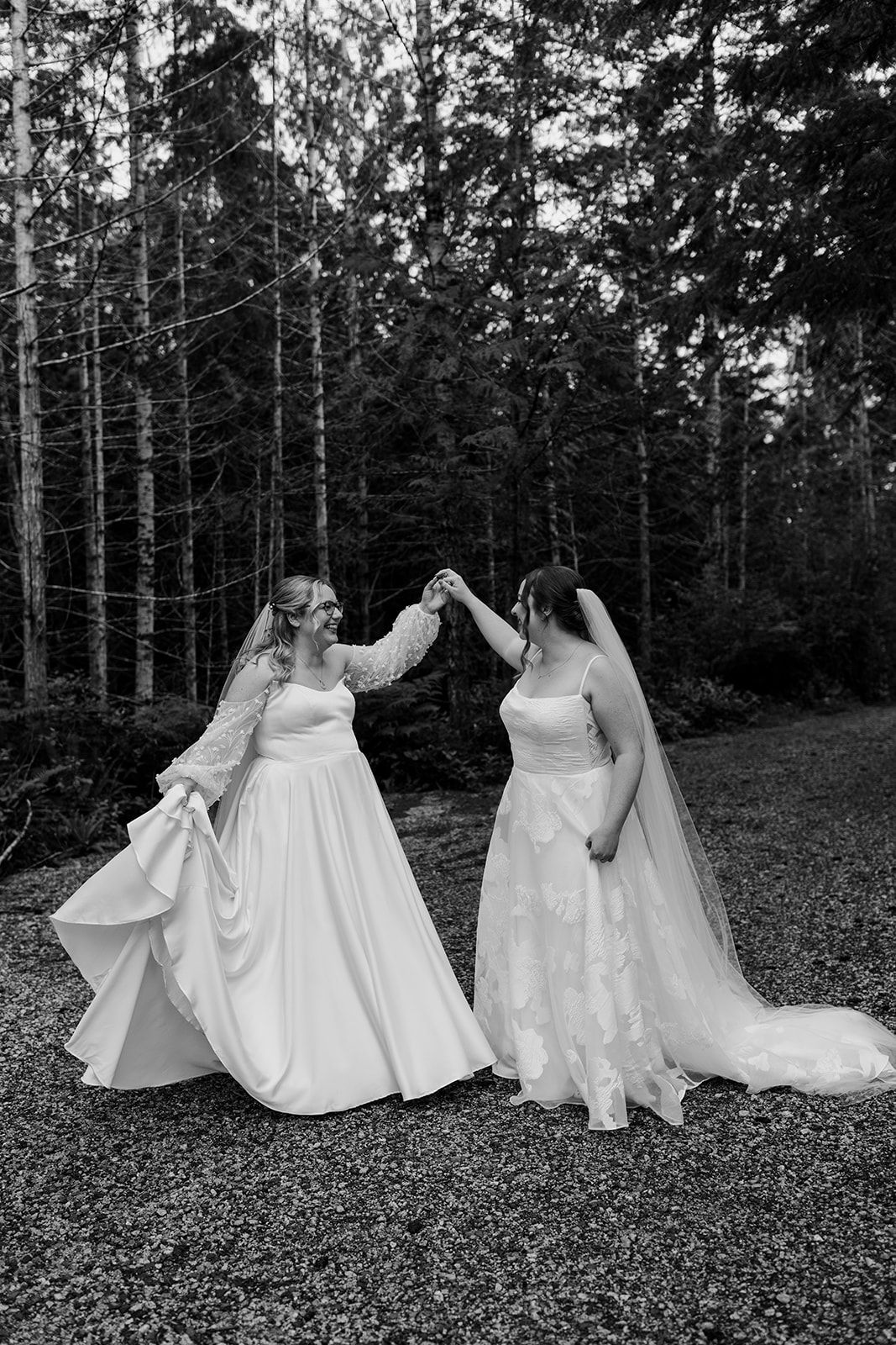 Black and white photo of two brides in elegant dresses dancing in a forest clearing.