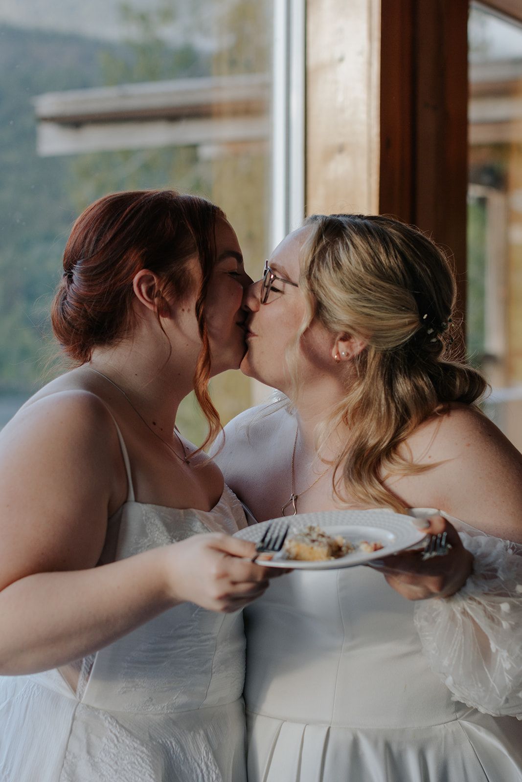 Couple in white dresses sharing cake during their wedding reception.