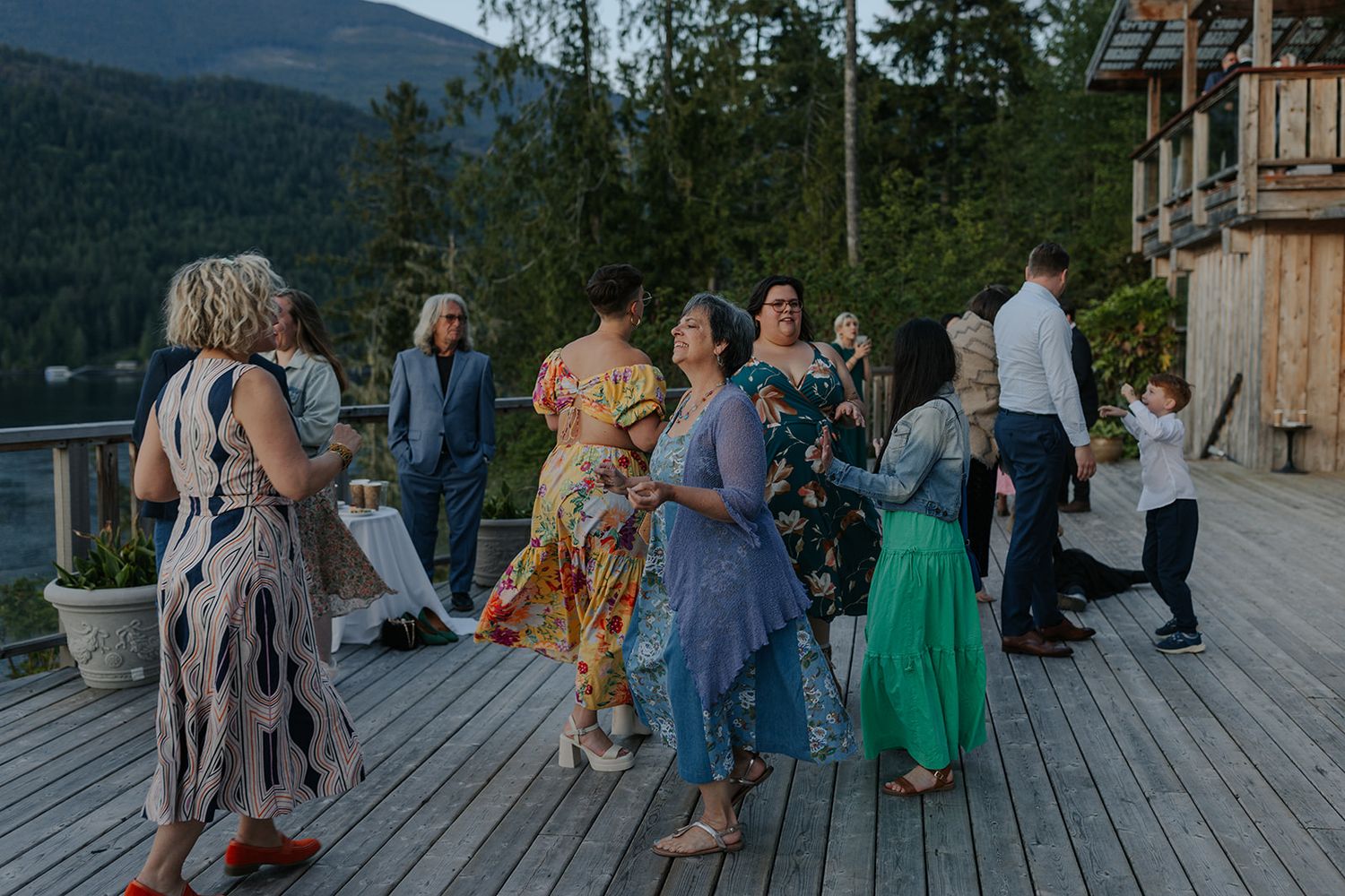 Wedding guests dance in colorful dresses on a deck with mountain views at dusk.