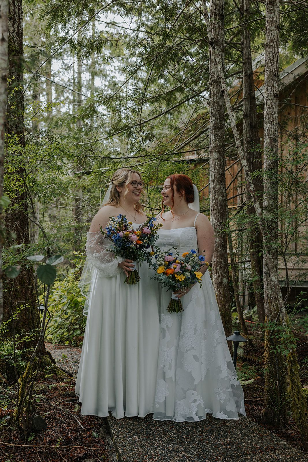 Two brides in white dresses share a joyful moment while holding colorful bouquets in a misty forest setting.