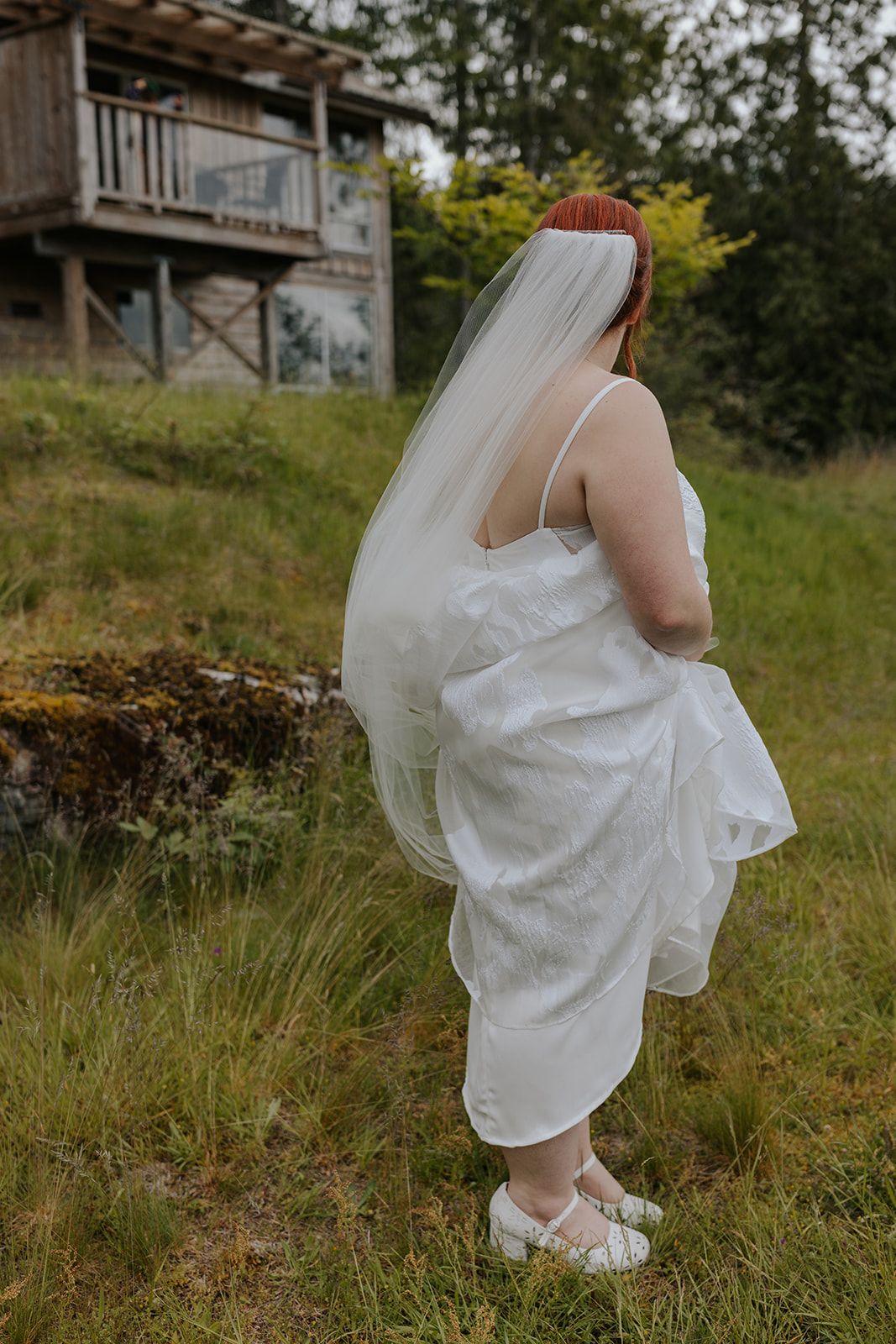 A bride in a white dress and veil stands on a grassy hillside near a wooden deck structure.