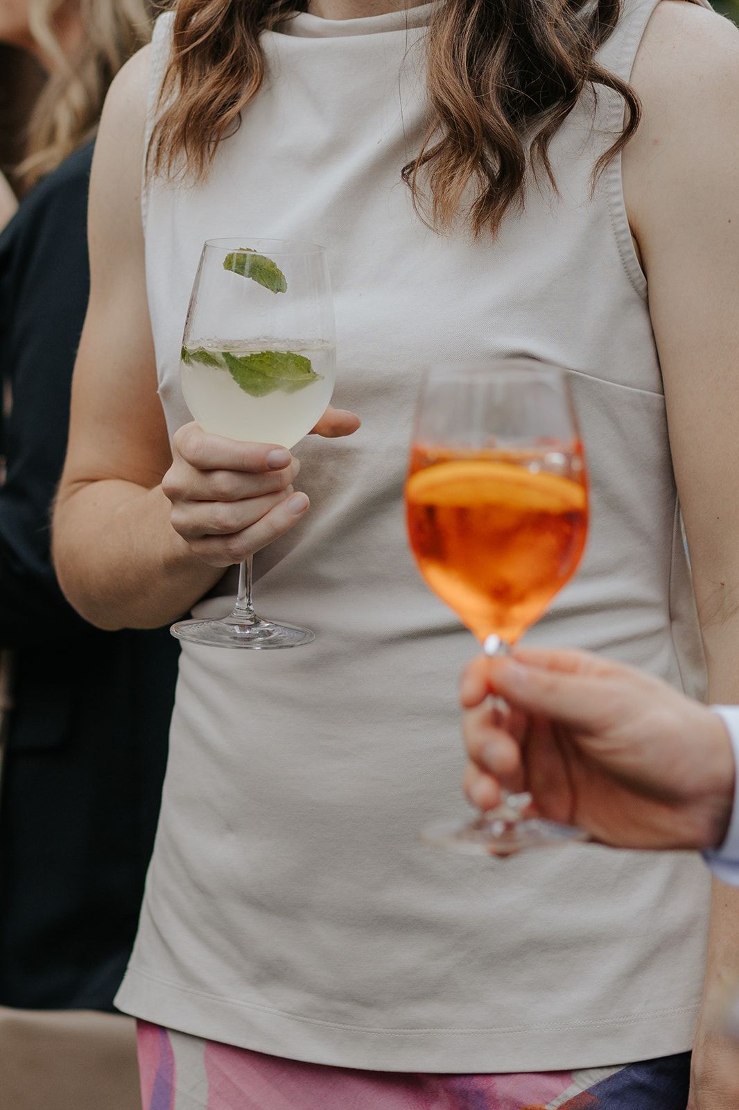 People holding cocktail glasses with orange-colored drinks garnished with fresh herbs.