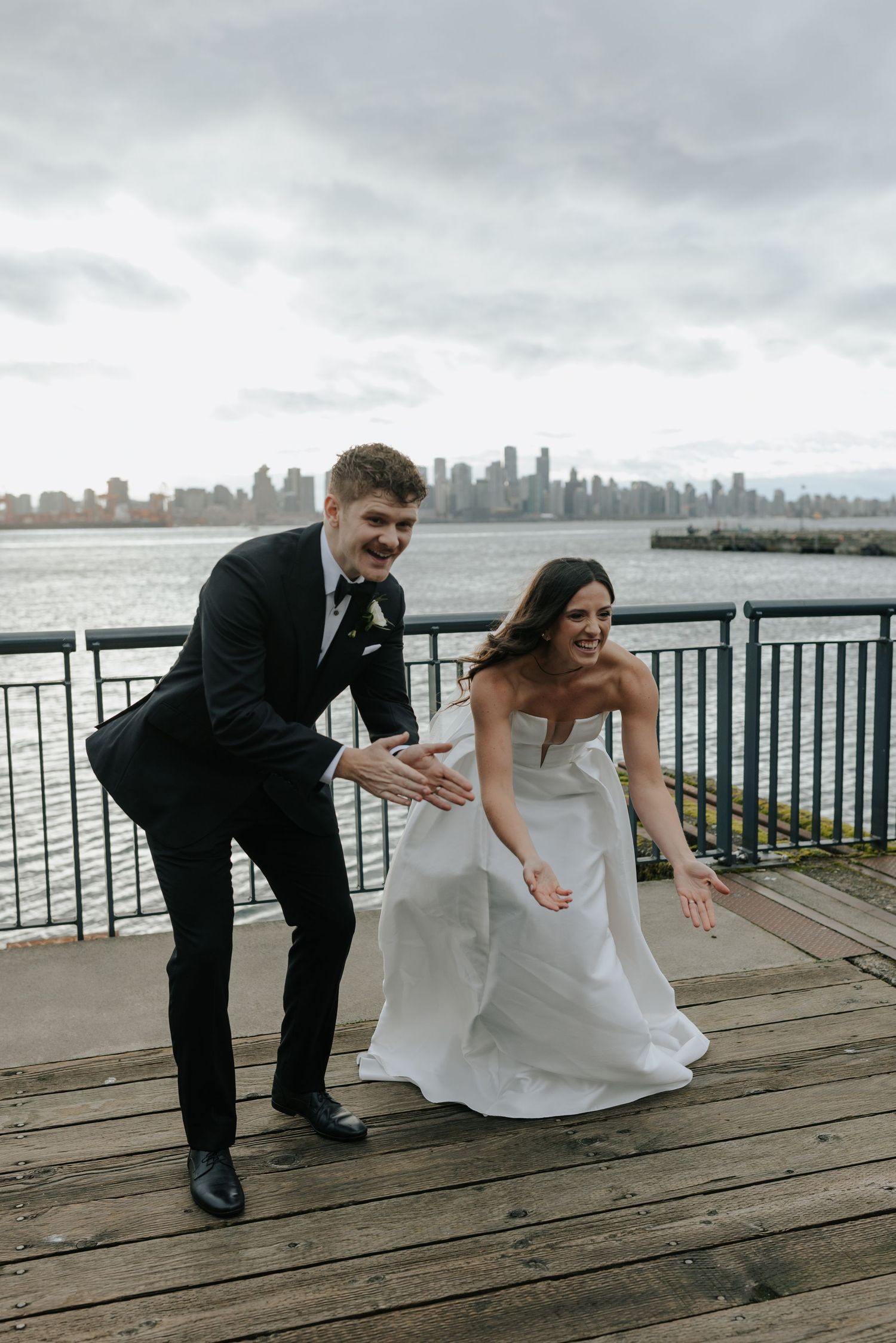 A couple bends down laughing together on a wooden pier with a city skyline visible across the water.