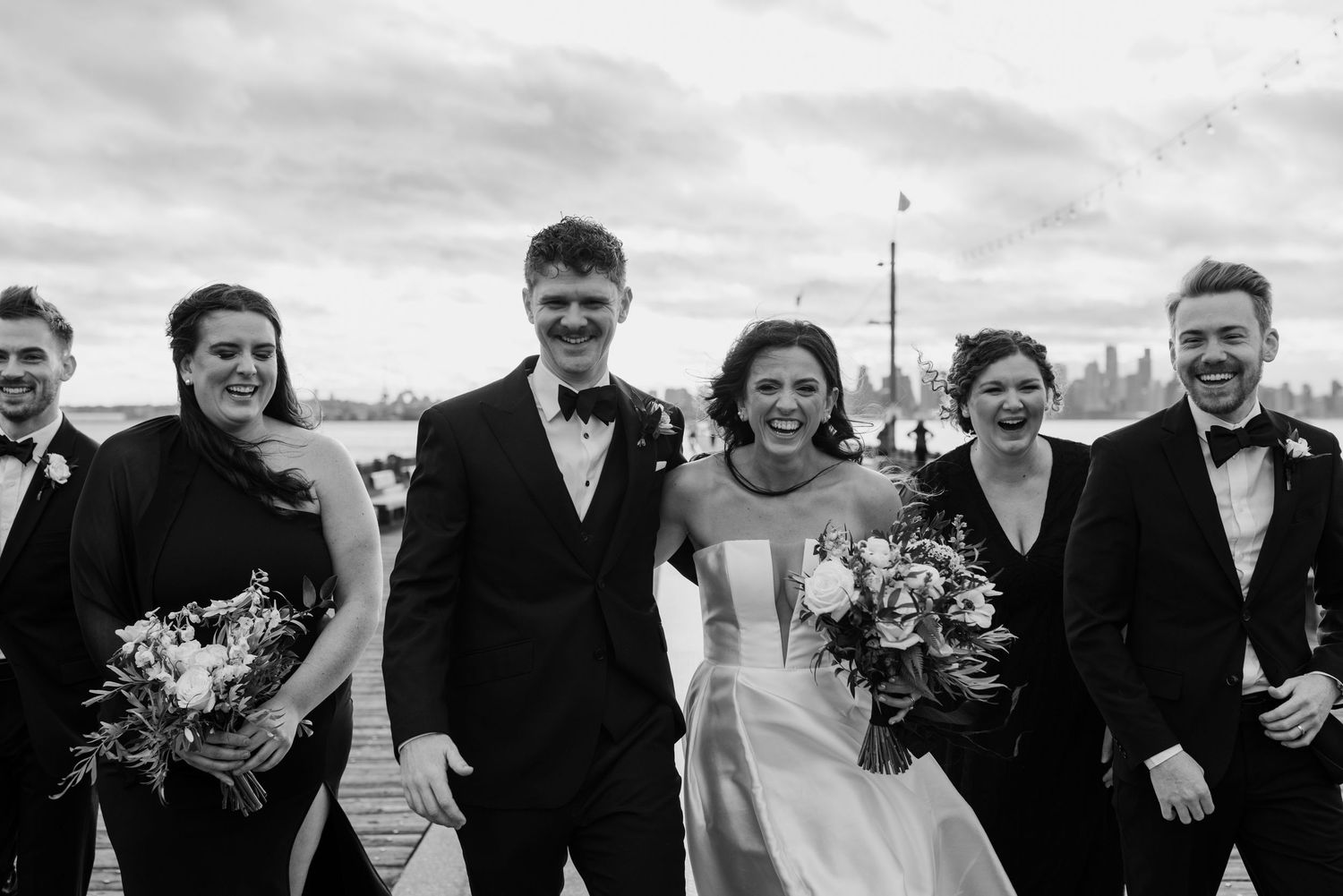 A wedding party in formal attire poses together on a cloudy day by the water.
