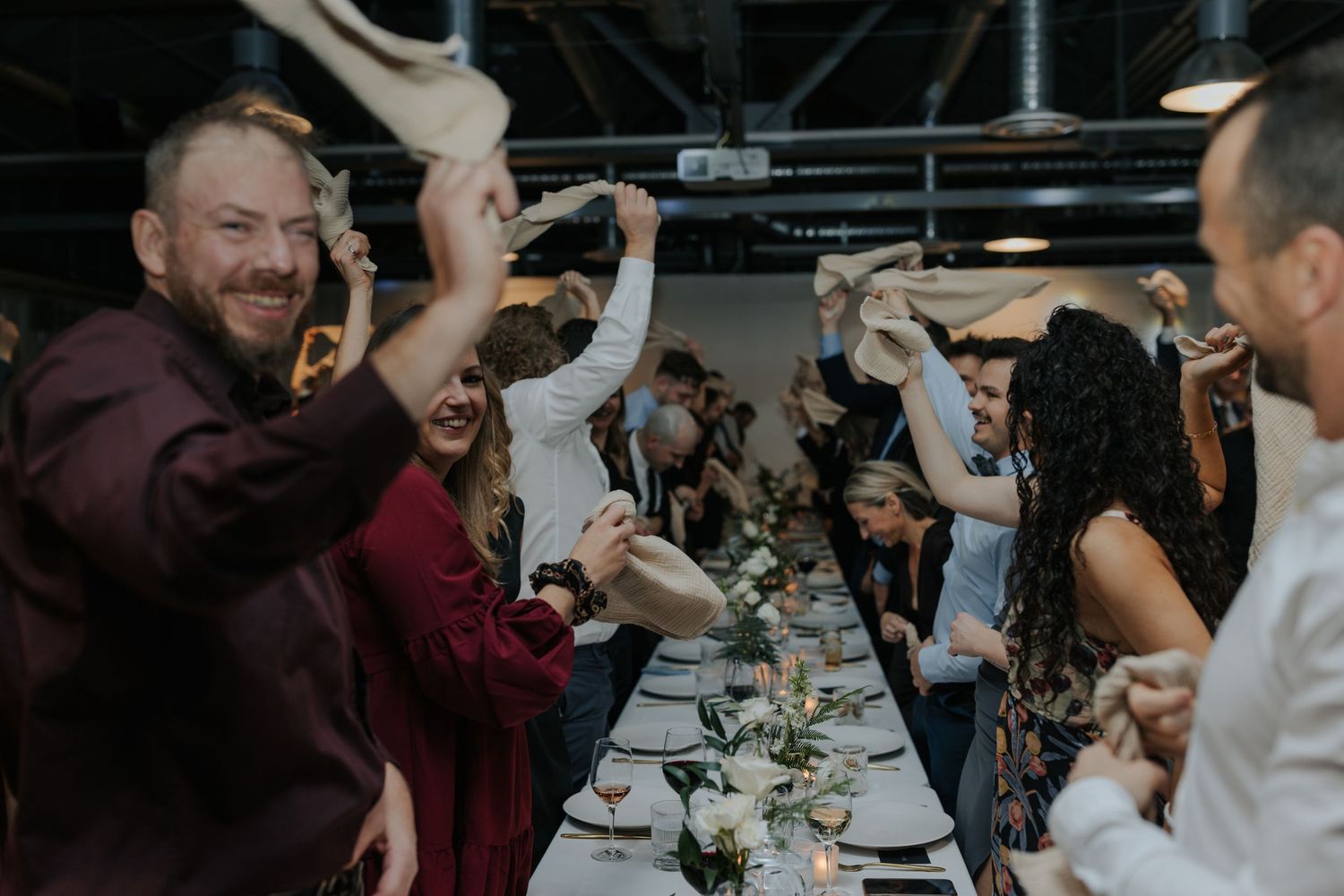 Wedding reception guests celebrate and dance under industrial lighting.