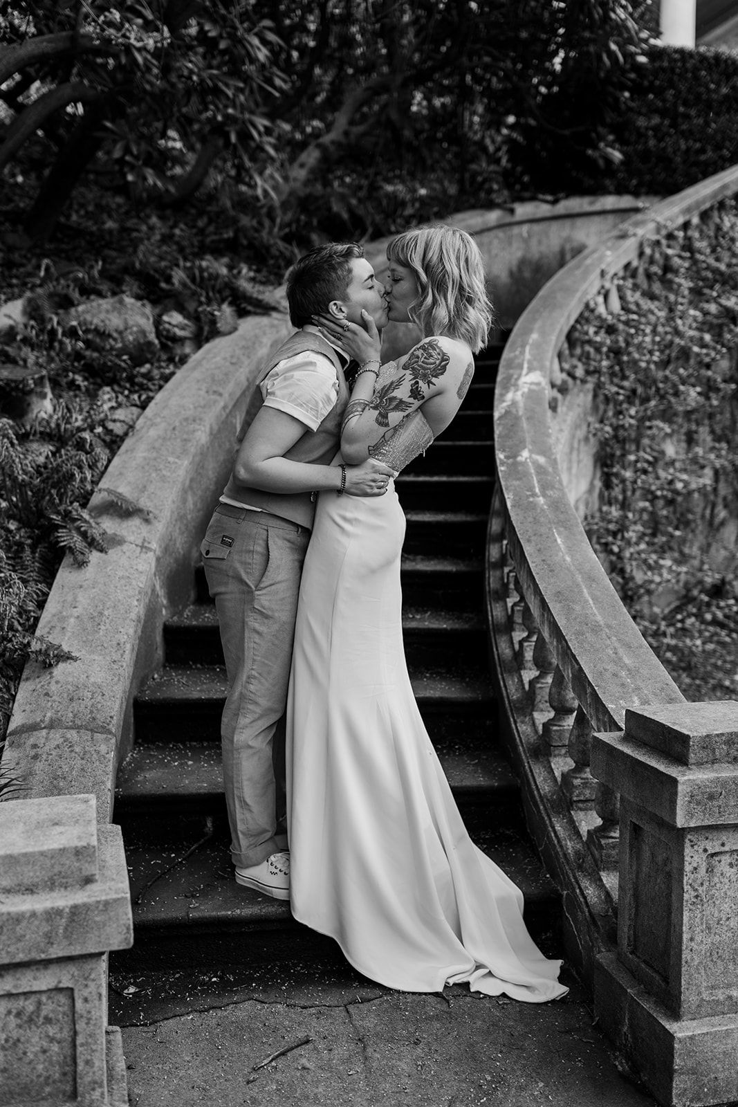 Romantic black and white photo of a couple embracing on stone steps surrounded by foliage.