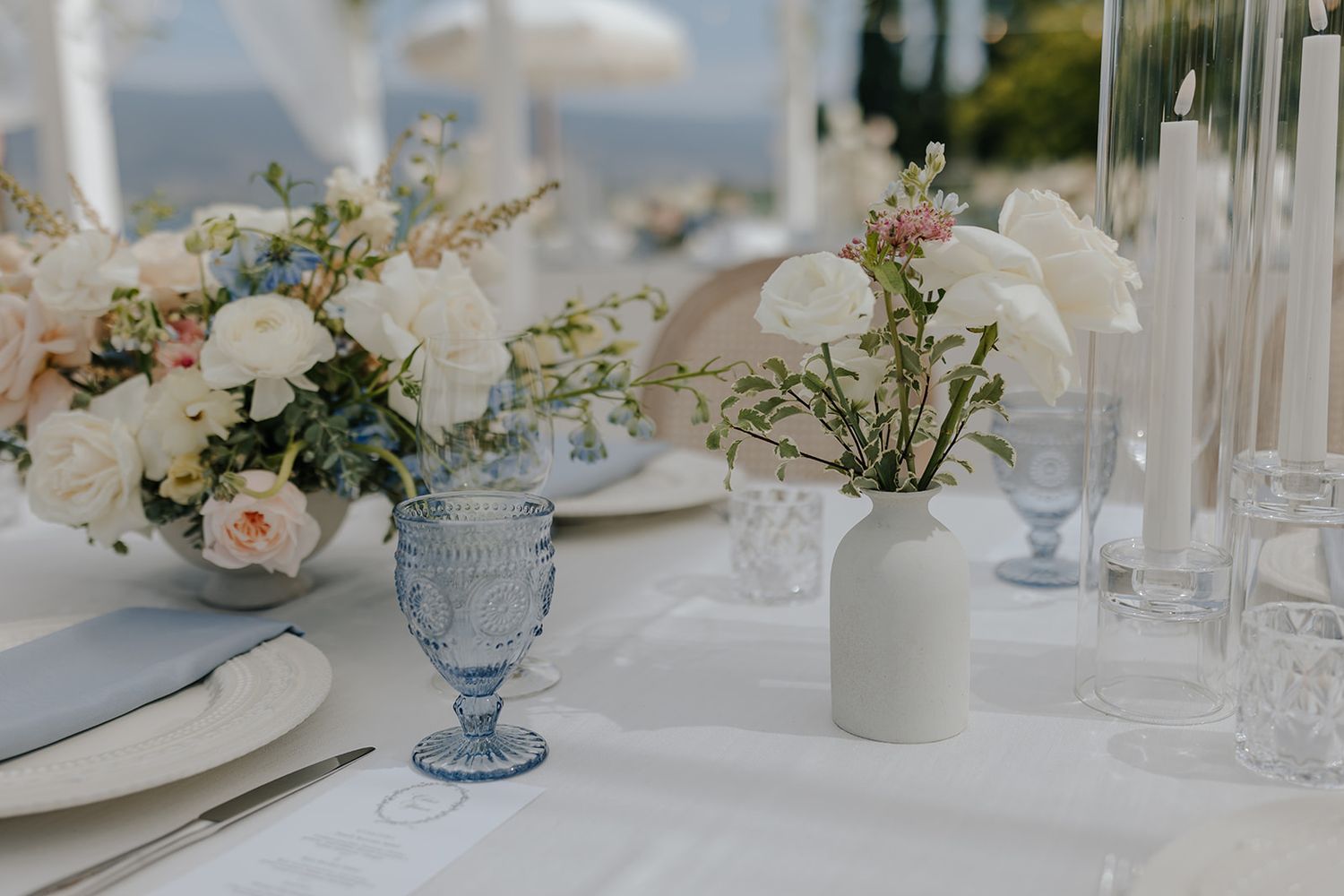 Elegant wedding reception table settings with blue glass vases and delicate floral centerpieces on white tablecloths.