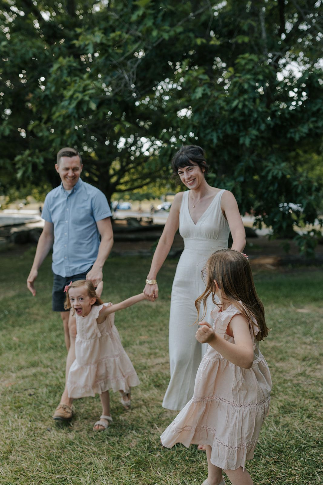 Family members in light summer clothing dancing together on grass in a park.