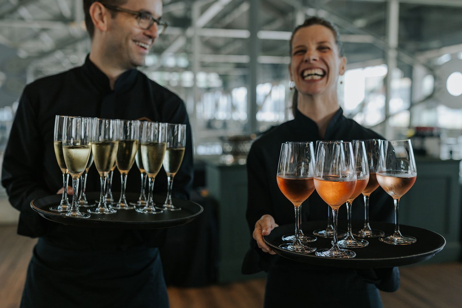 Two servers carrying trays of champagne and wine glasses in an industrial venue.