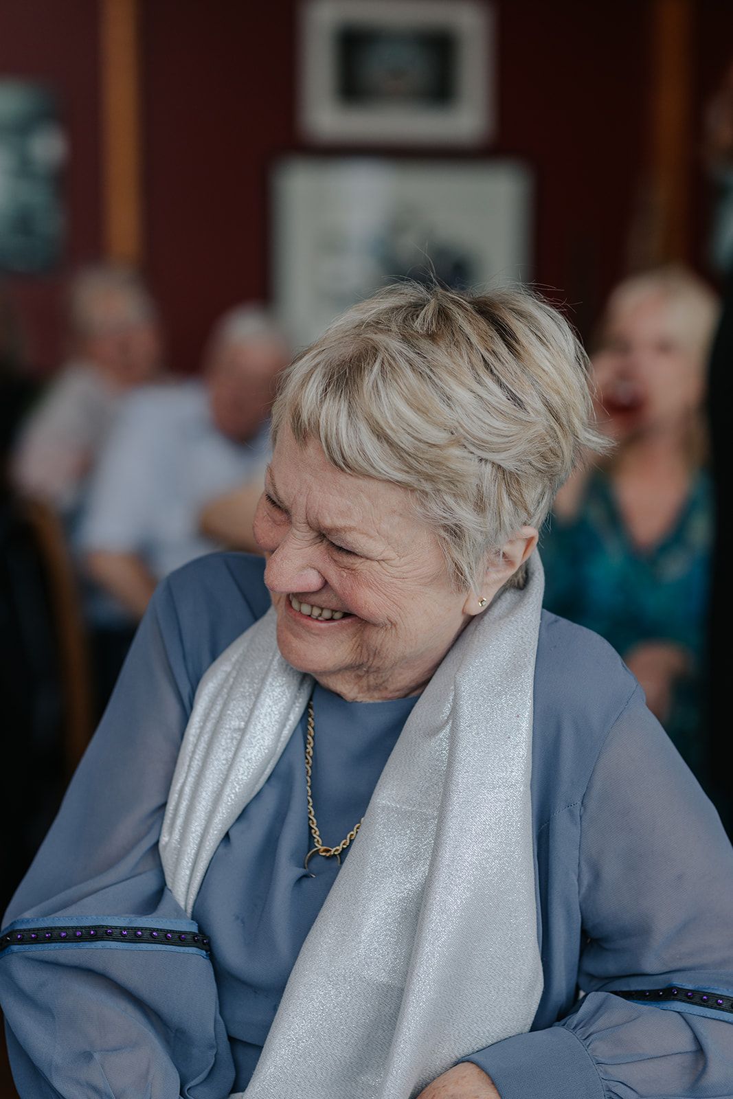 Senior guest wearing blue sweater and white scarf laughs heartily at celebration.