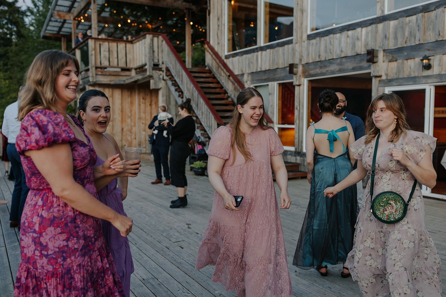 Wedding guests in formal dresses chat during outdoor reception on wooden deck.