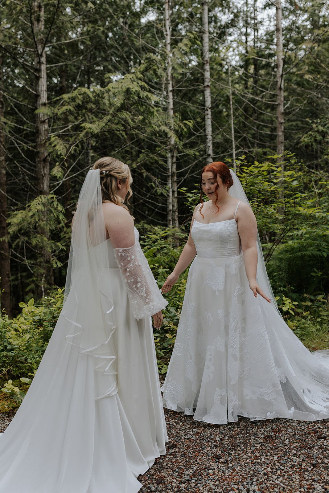 Two brides in white wedding dresses stand together on a forest path.