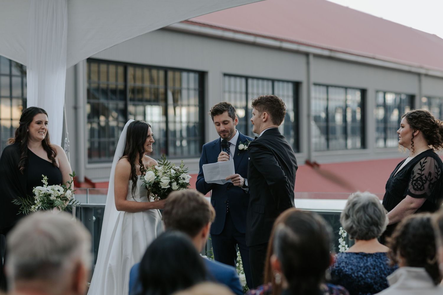 Wedding ceremony guests gather indoors with large windows and industrial architecture.