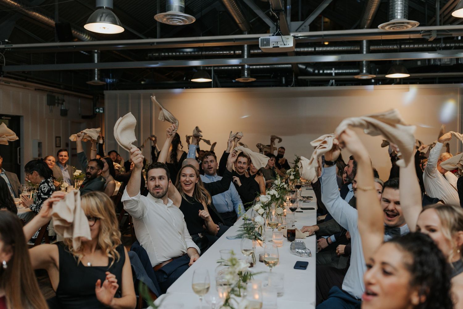 Wedding reception guests celebrating with arms raised at long banquet table.