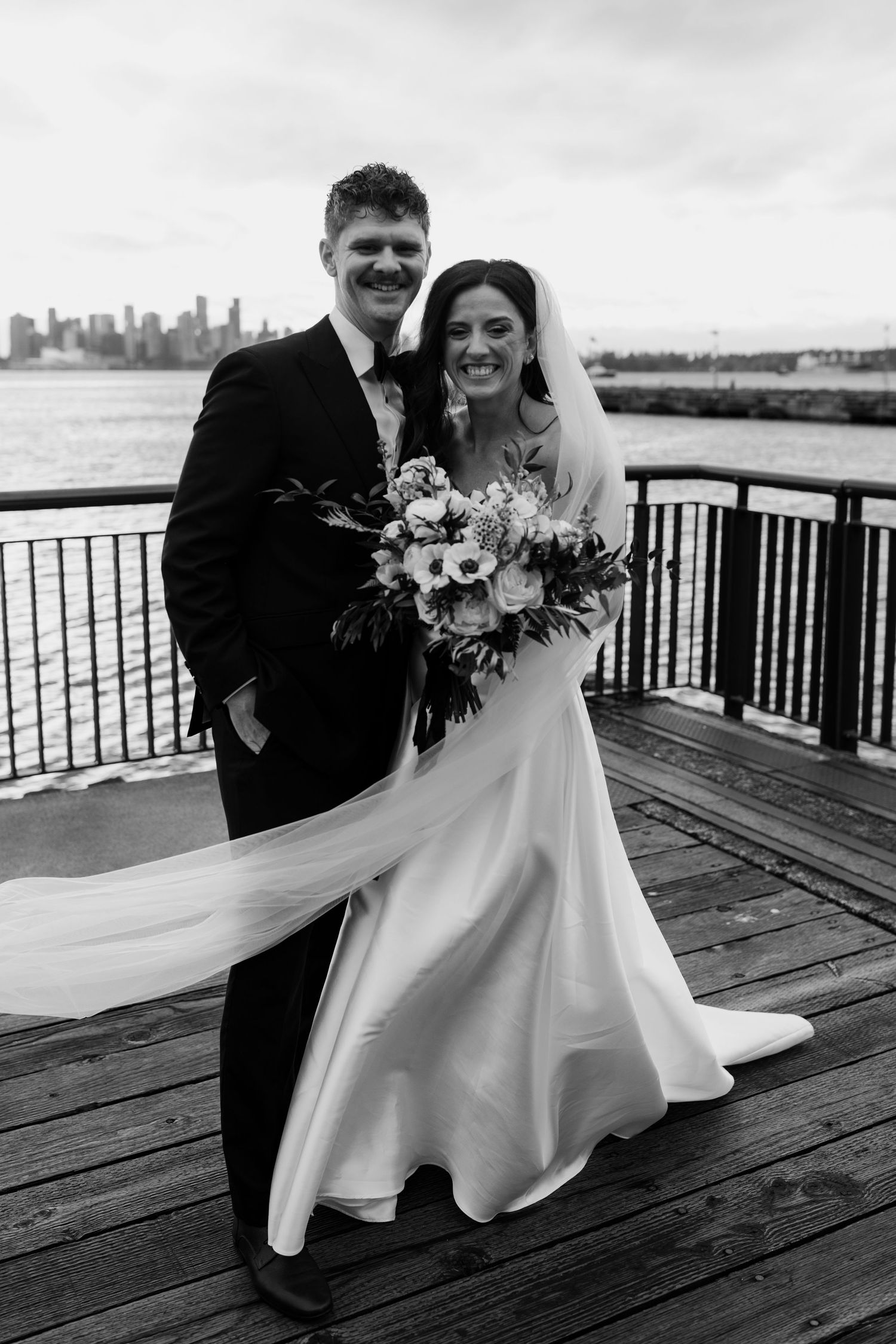 Black and white wedding portraits of a couple embracing on a wooden deck overlooking a waterfront cityscape.