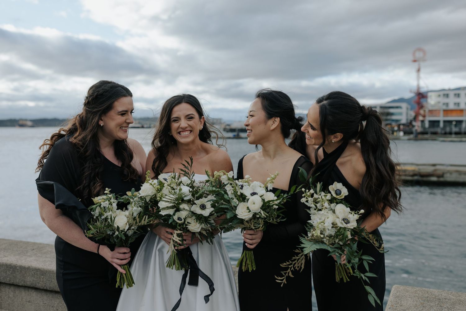 Bridesmaids in black dresses laugh together while holding floral bouquets by the waterfront.