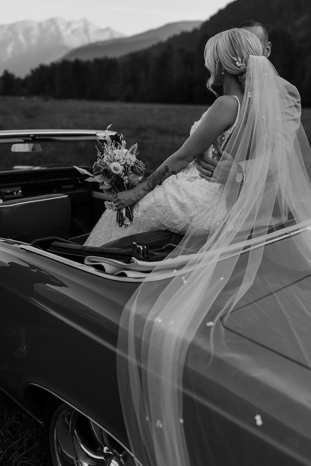 Black and white photo of bridal veil flowing over vintage car with mountain scenery.