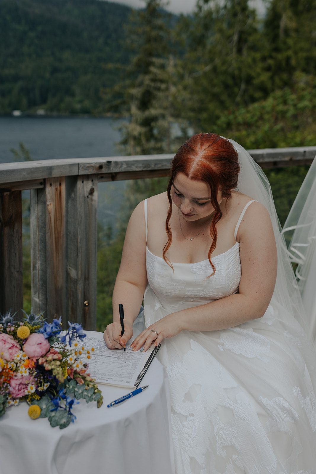 Two people in white wedding dresses sign documents with colorful flowers on the table.
