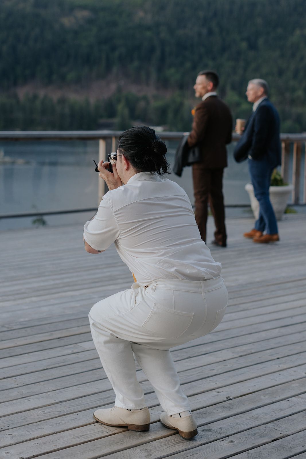 Photographer in white outfit crouches to take photos on wooden deck by mountain lake.