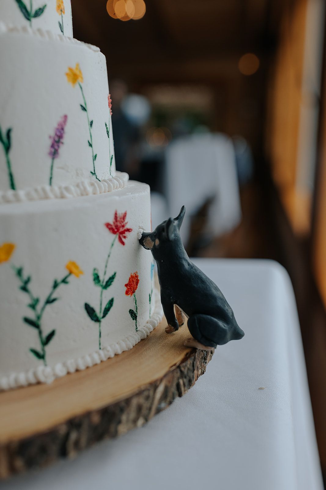White tiered wedding cake decorated with delicate floral designs and a small black dog figurine on a wood slice base.
