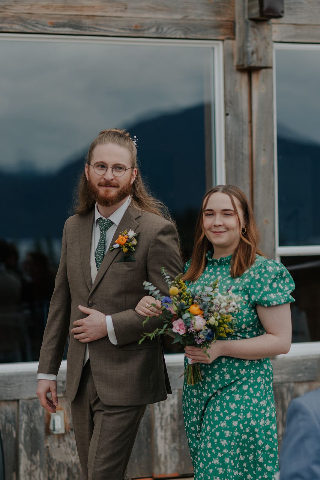 Couple in brown suit and green floral dress pose with bouquet at mountain venue.