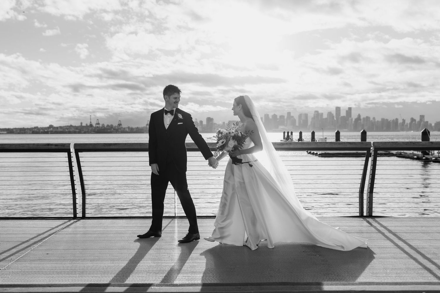 Romantic wedding couple silhouettes embrace on a pier at sunset with water and cityscape views in the background.