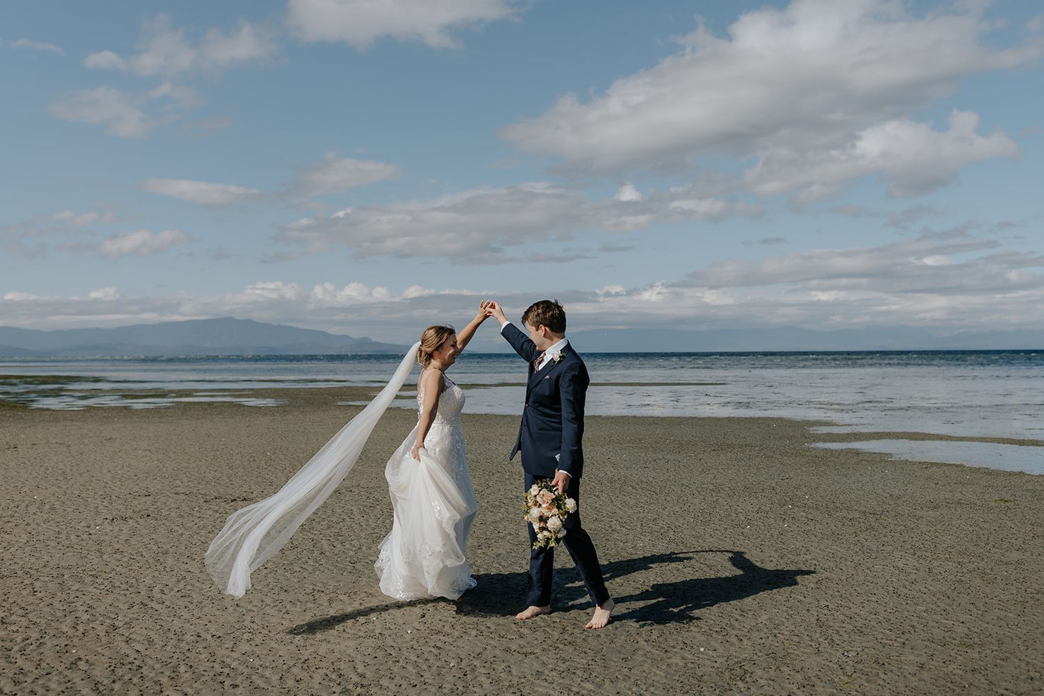 Couple poses for romantic wedding photos on a scenic beach with mountains and dramatic clouds in the background.