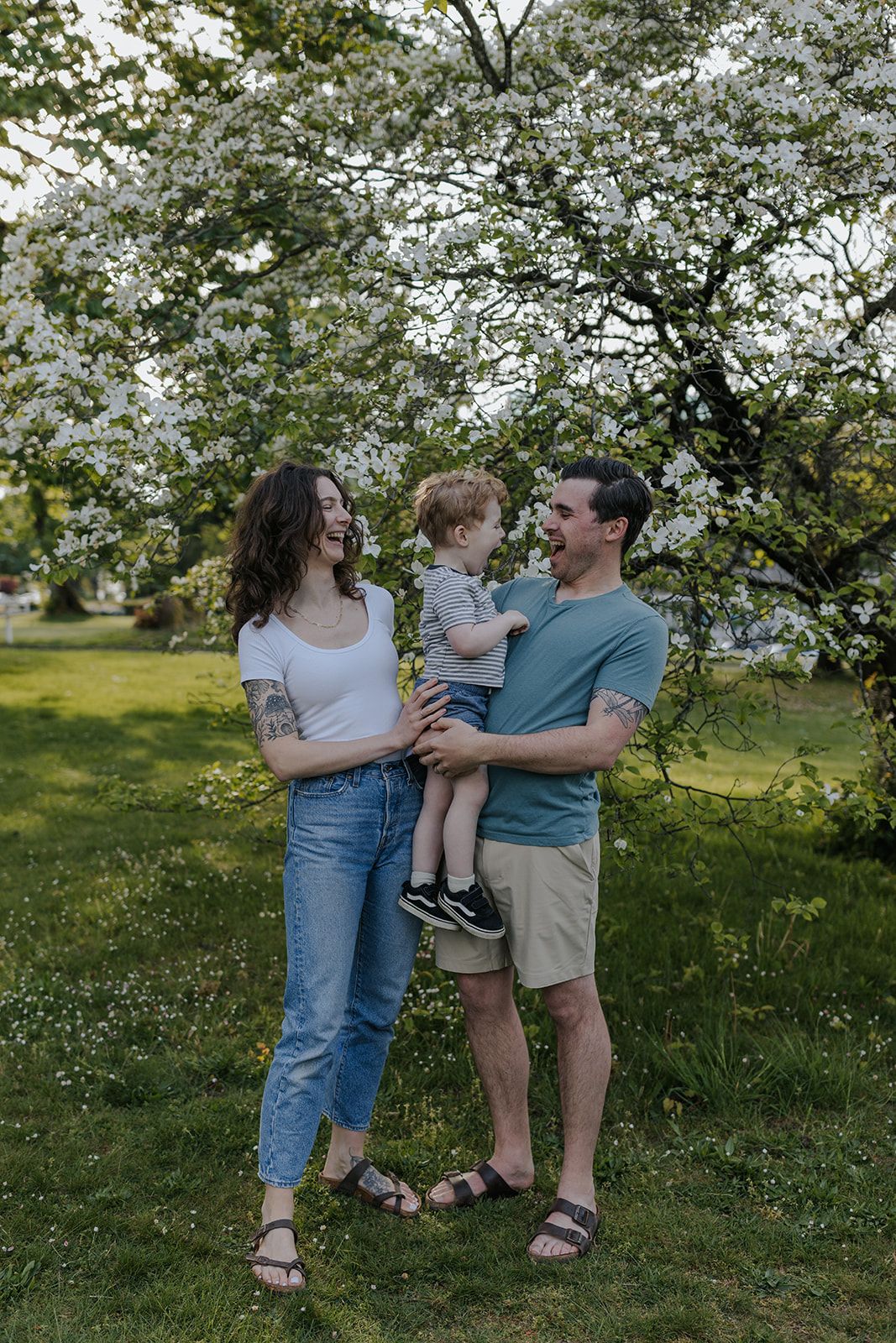 A happy young family with a toddler poses outdoors near a flowering tree on a sunny day.