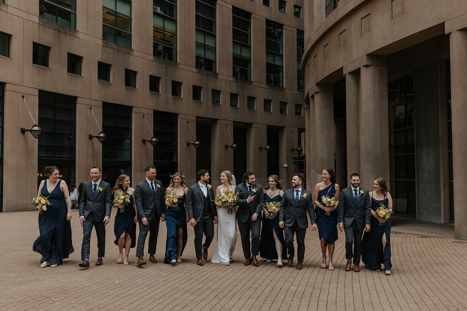 Large group photo of wedding party members lined up against modern building architecture.