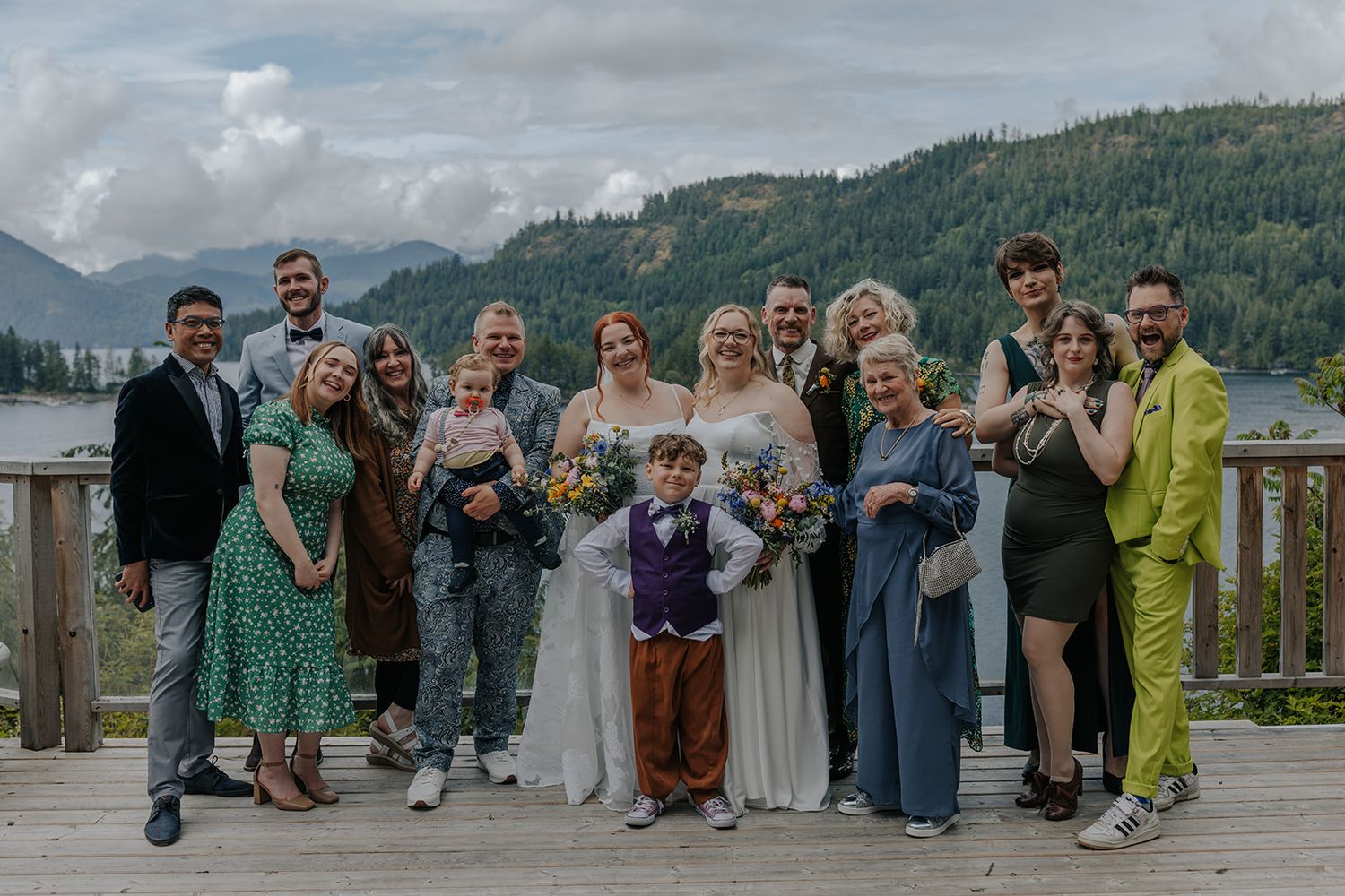 Group photo of wedding attendees in formal attire on deck with mountain and lake views.