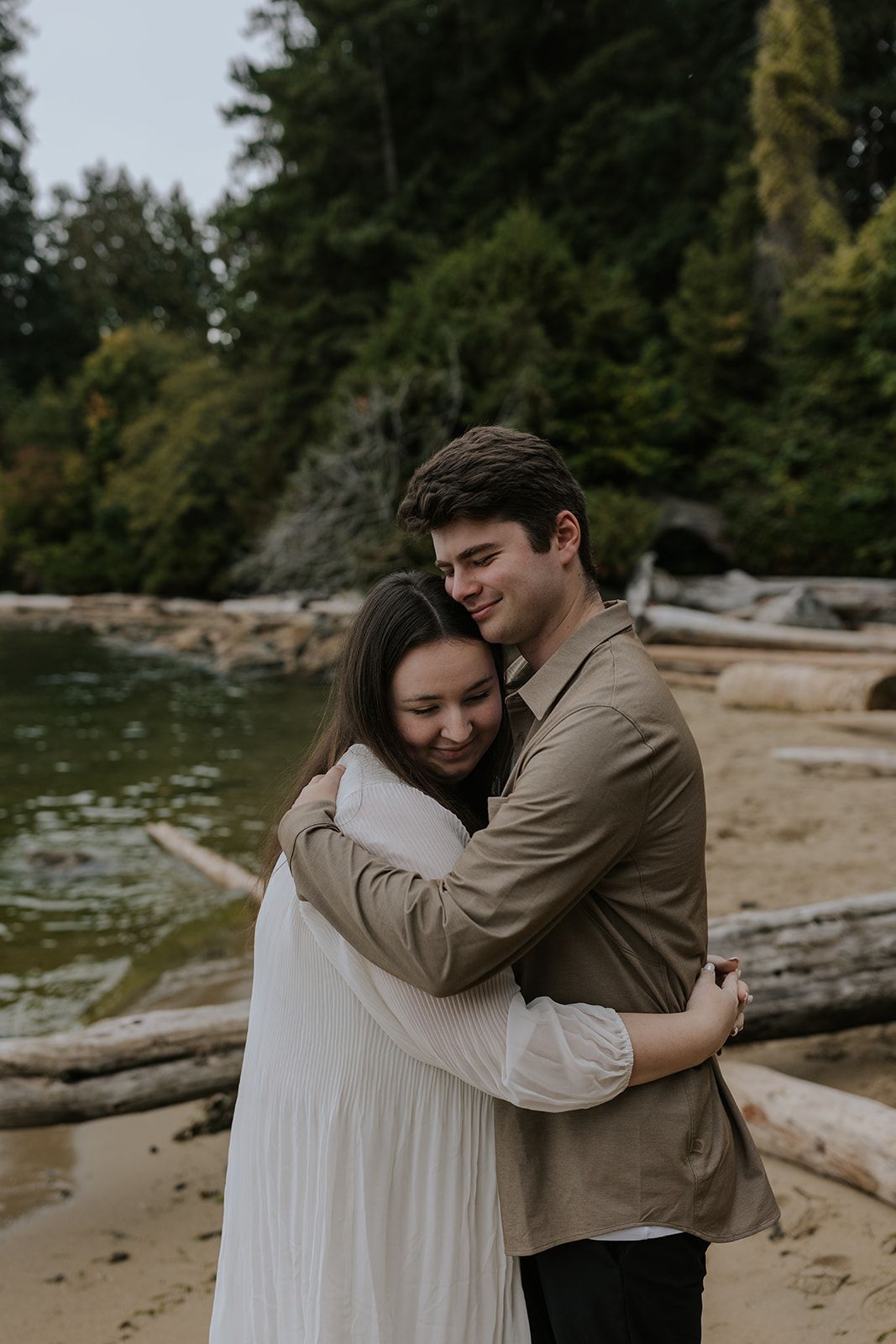 Couple embracing on a beach with driftwood and forest in background.