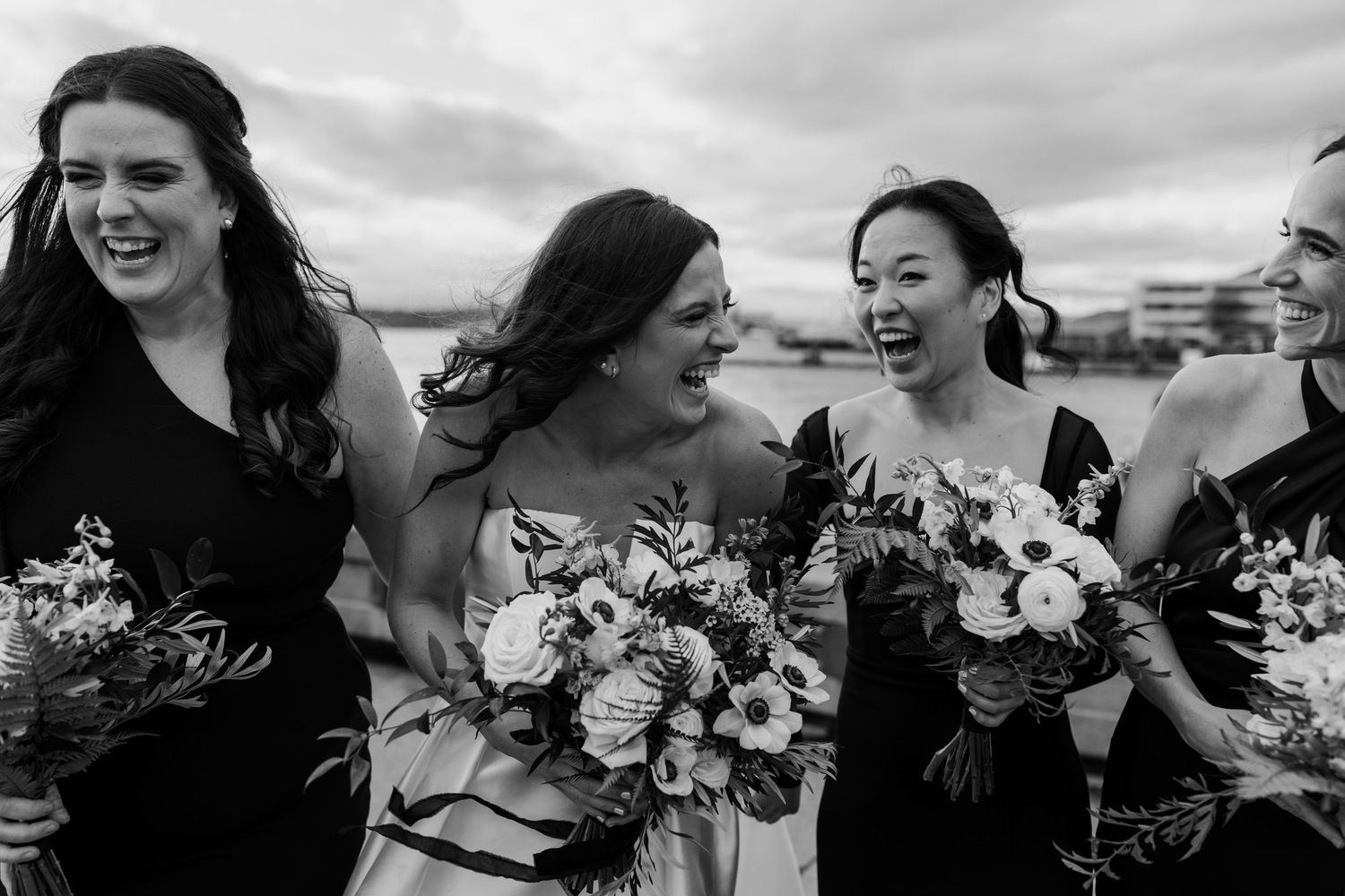 A candid black and white photo showing a group of bridesmaids laughing together while holding floral bouquets.