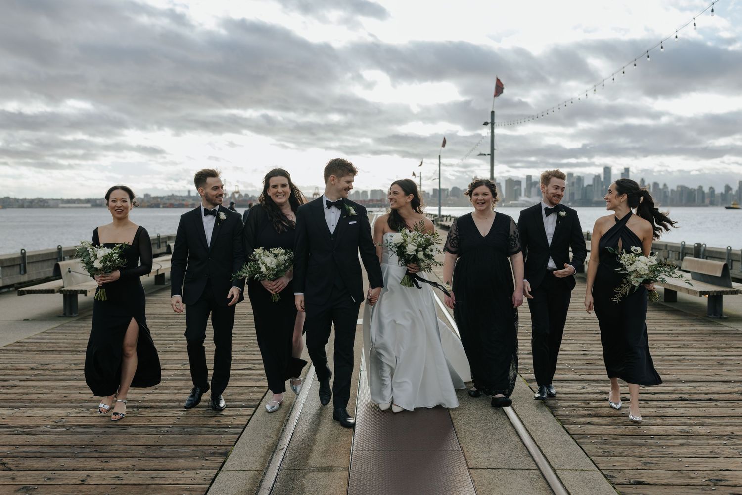 Wedding party members lined up along a waterfront pier on an overcast day.