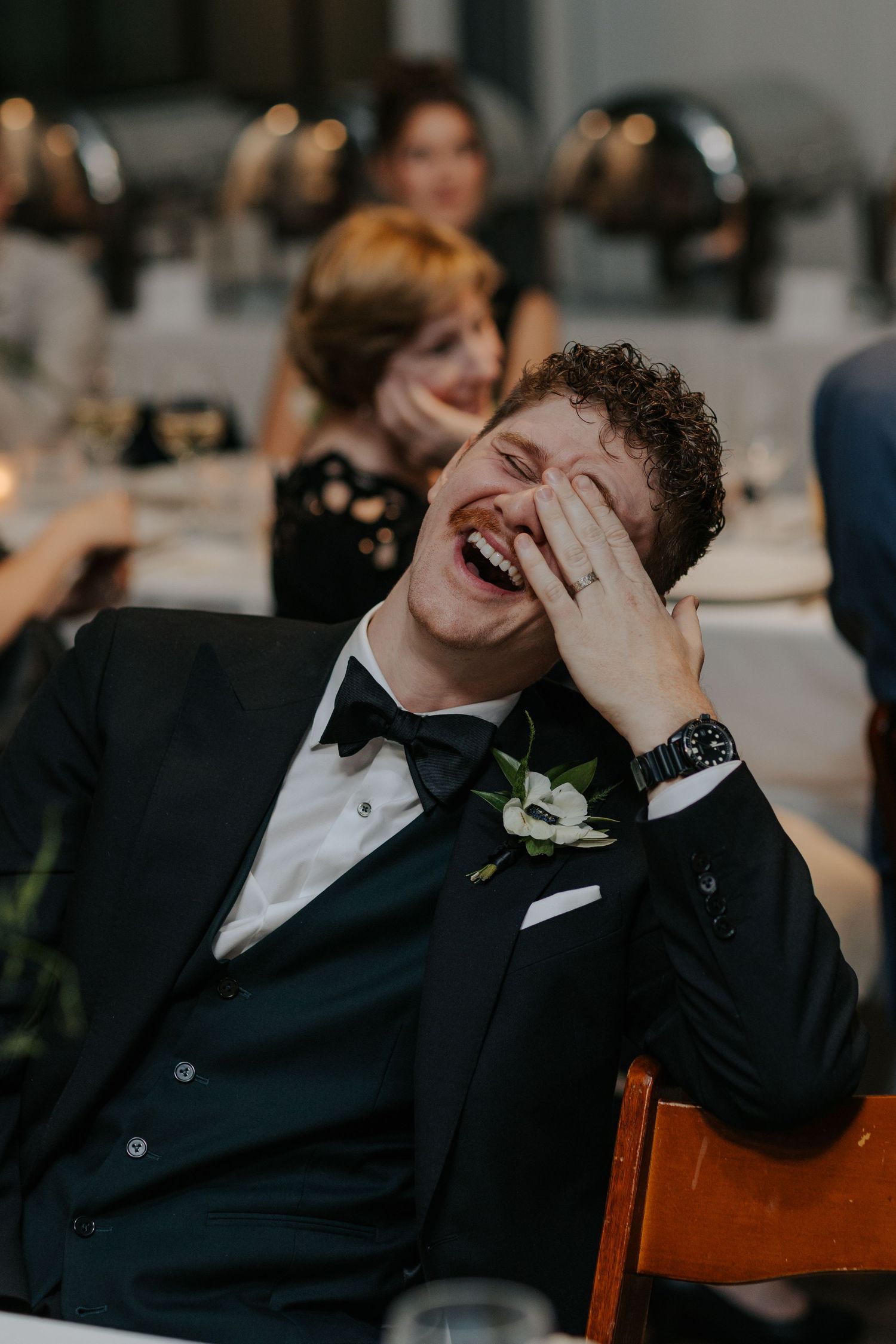 Wedding guest in formal attire laughing while seated at reception table.