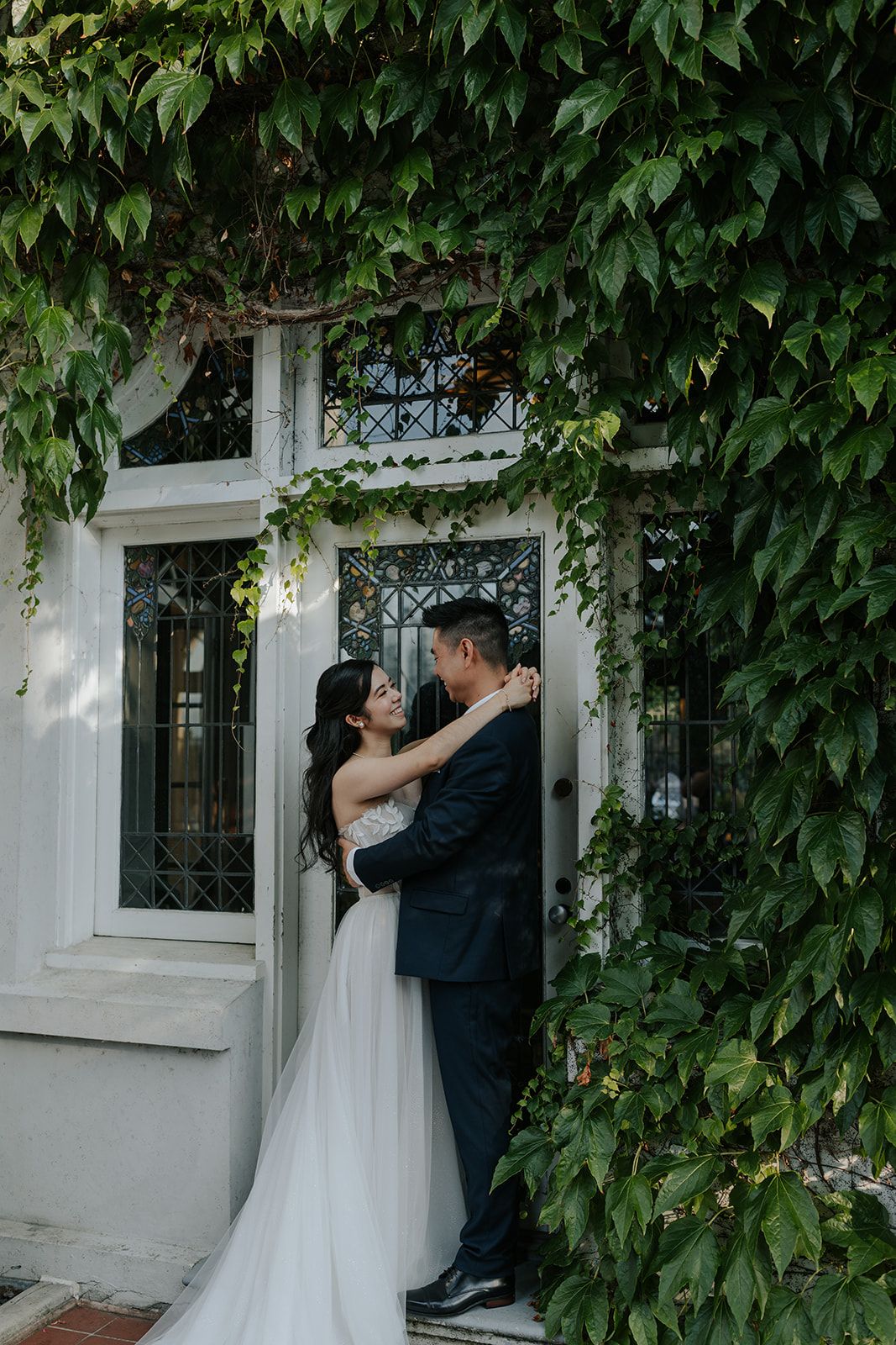 Romantic wedding portrait against ivy-covered wall with ornate window.