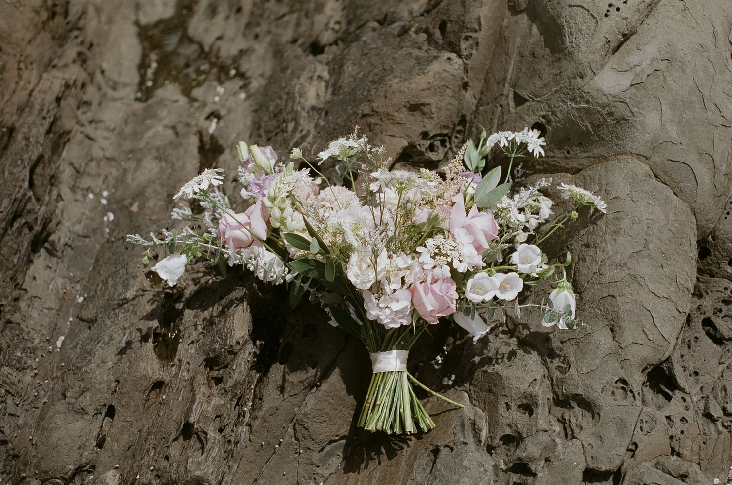 A delicate bridal bouquet with pink and white flowers rests on textured stone.
