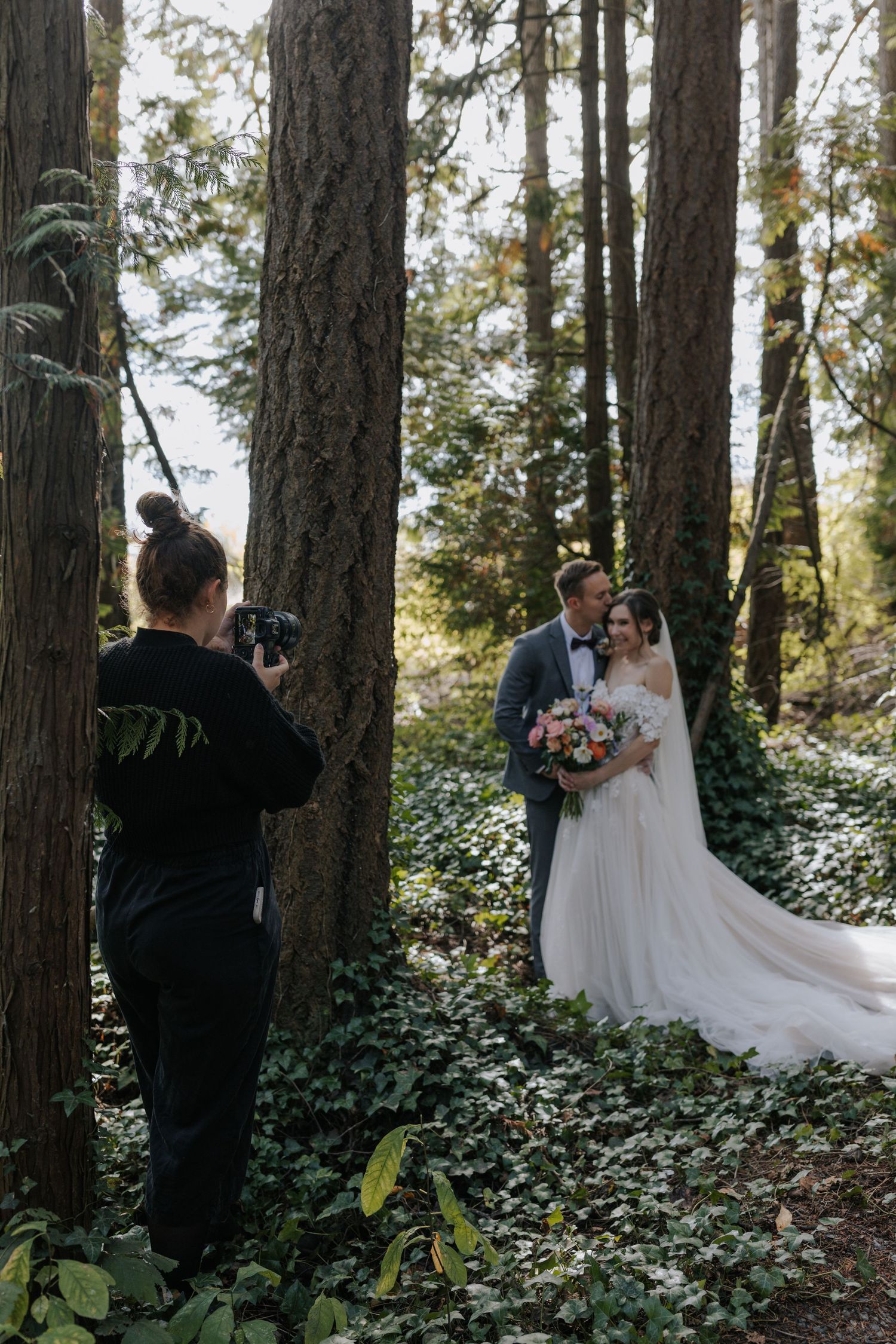 Wedding photographer taking pictures of couple among tall pine trees in forest setting.