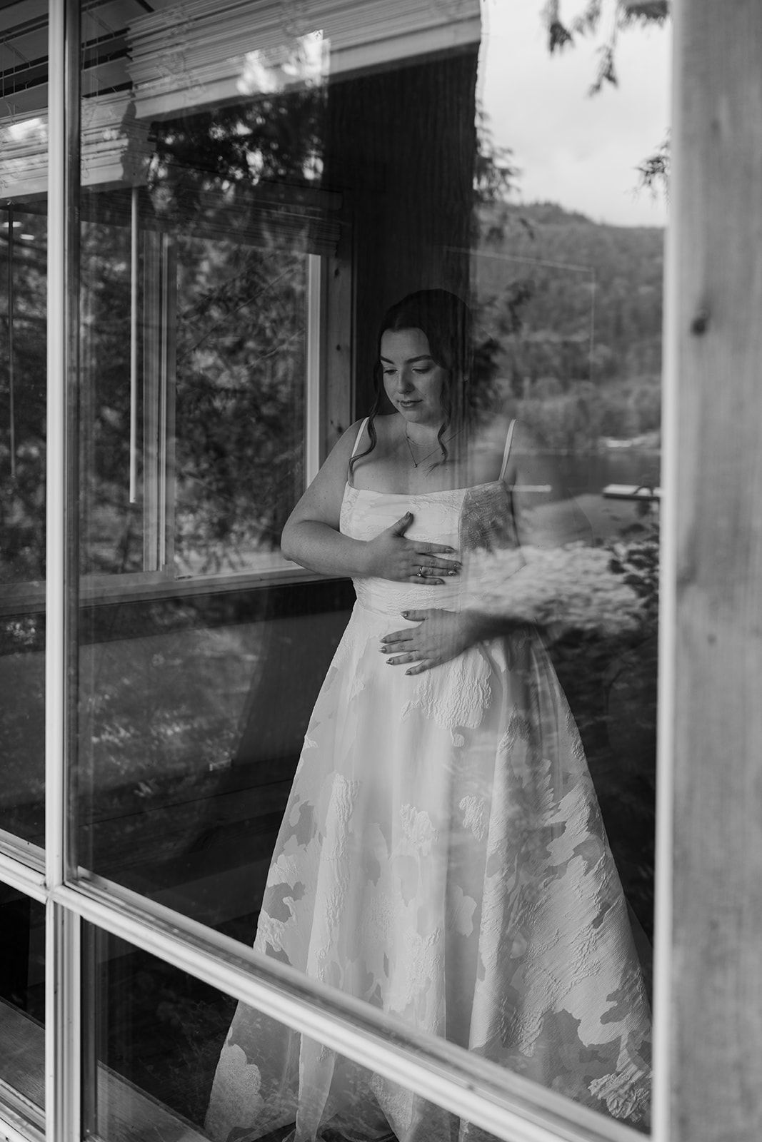 Artistic black and white photo of bride in white dress looking reflectively through window.