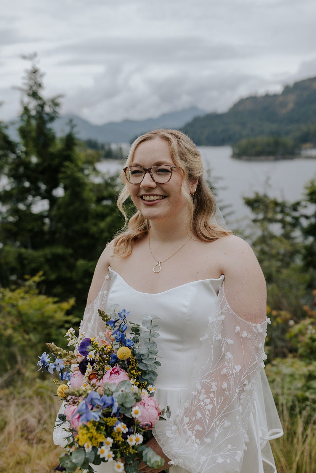 A bride in a white off-shoulder gown holds a colorful wildflower bouquet against a scenic mountain lake backdrop.