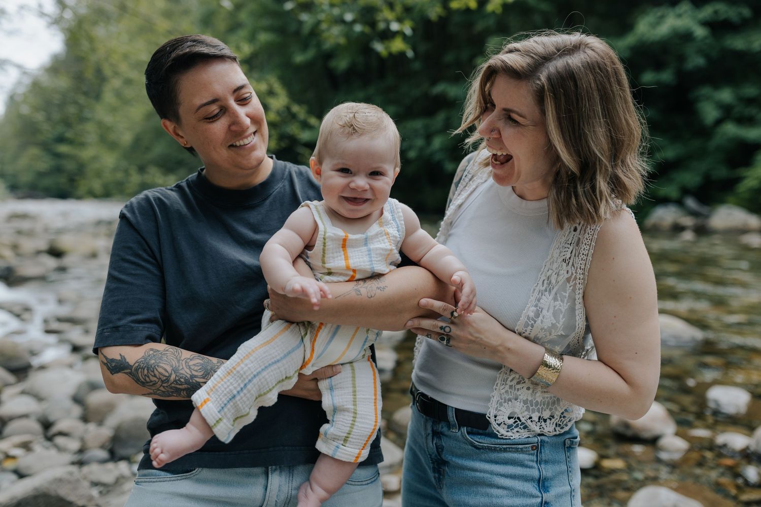 Two women smiling and holding a happy baby outdoors near a rocky river surrounded by lush green trees.