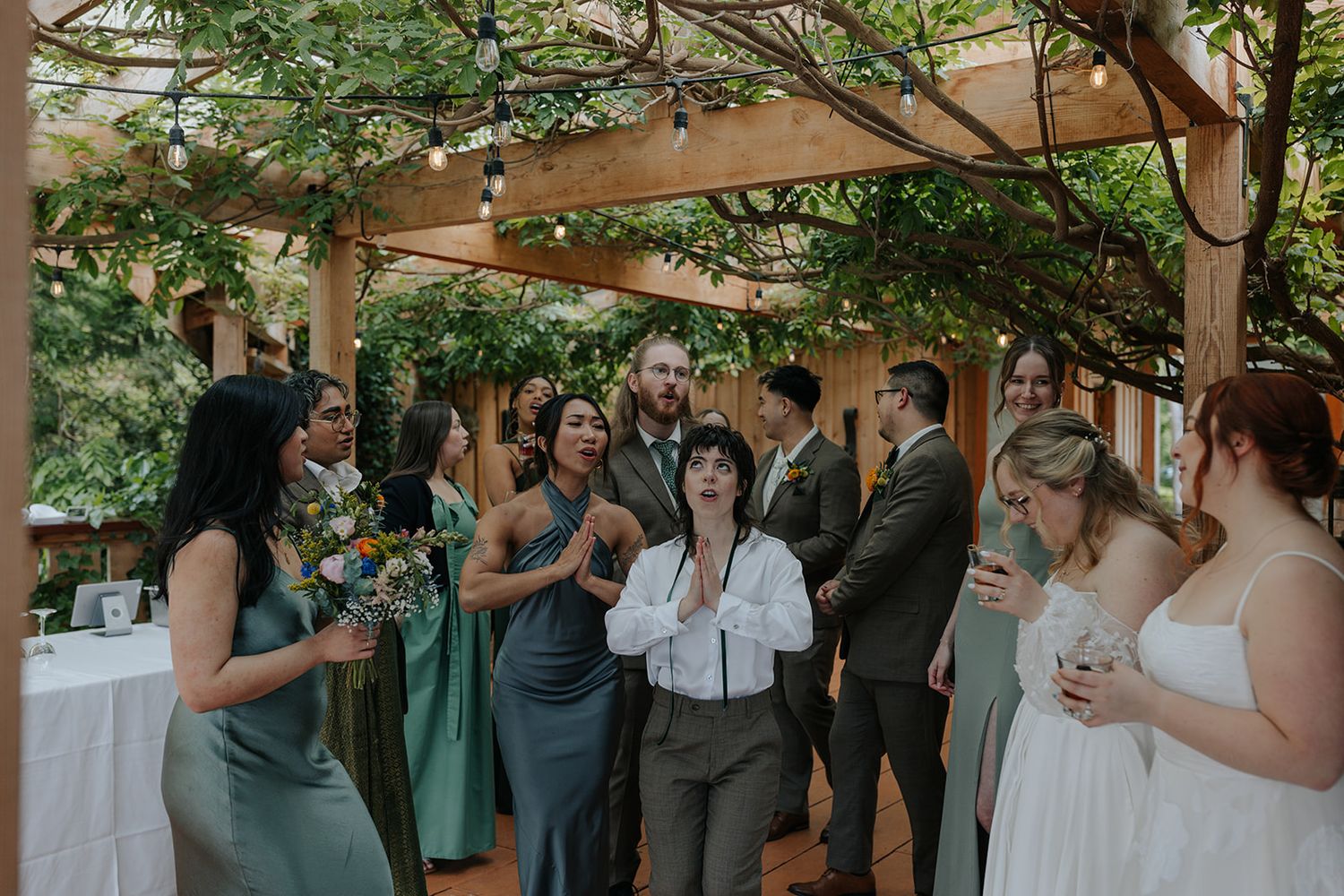 Wedding party gathered under a wooden pergola with greenery during an outdoor celebration.