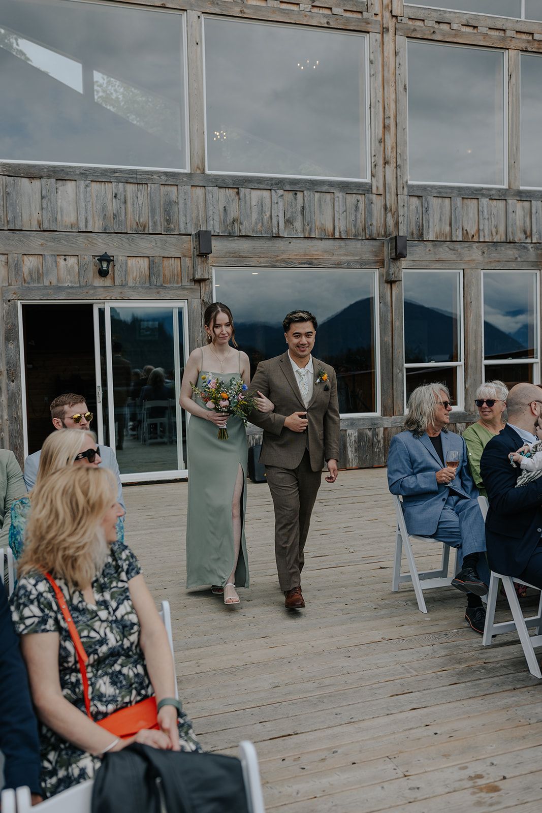 Wedding processional in rustic mountain venue with guests seated on white chairs.
