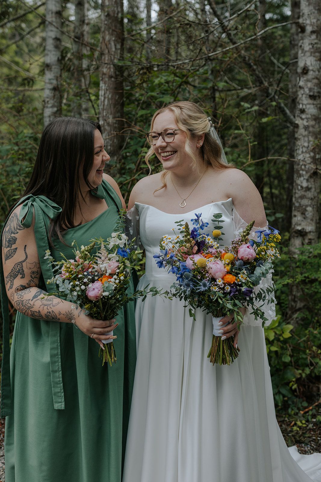 Wedding party members hold colorful bouquets featuring blue and orange flowers against a forest backdrop.