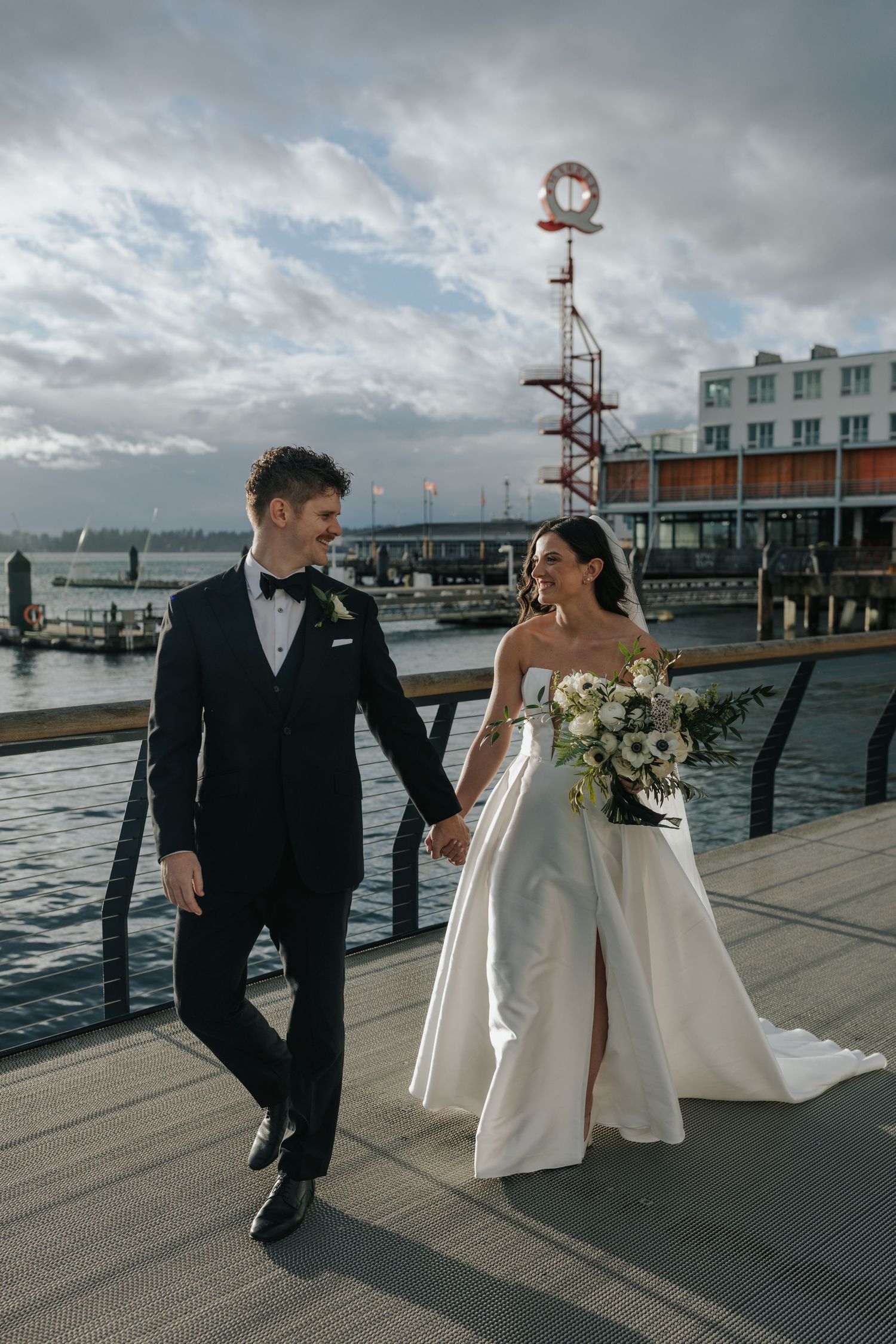 A wedding couple walks playfully along a waterfront pier with cloudy skies overhead.